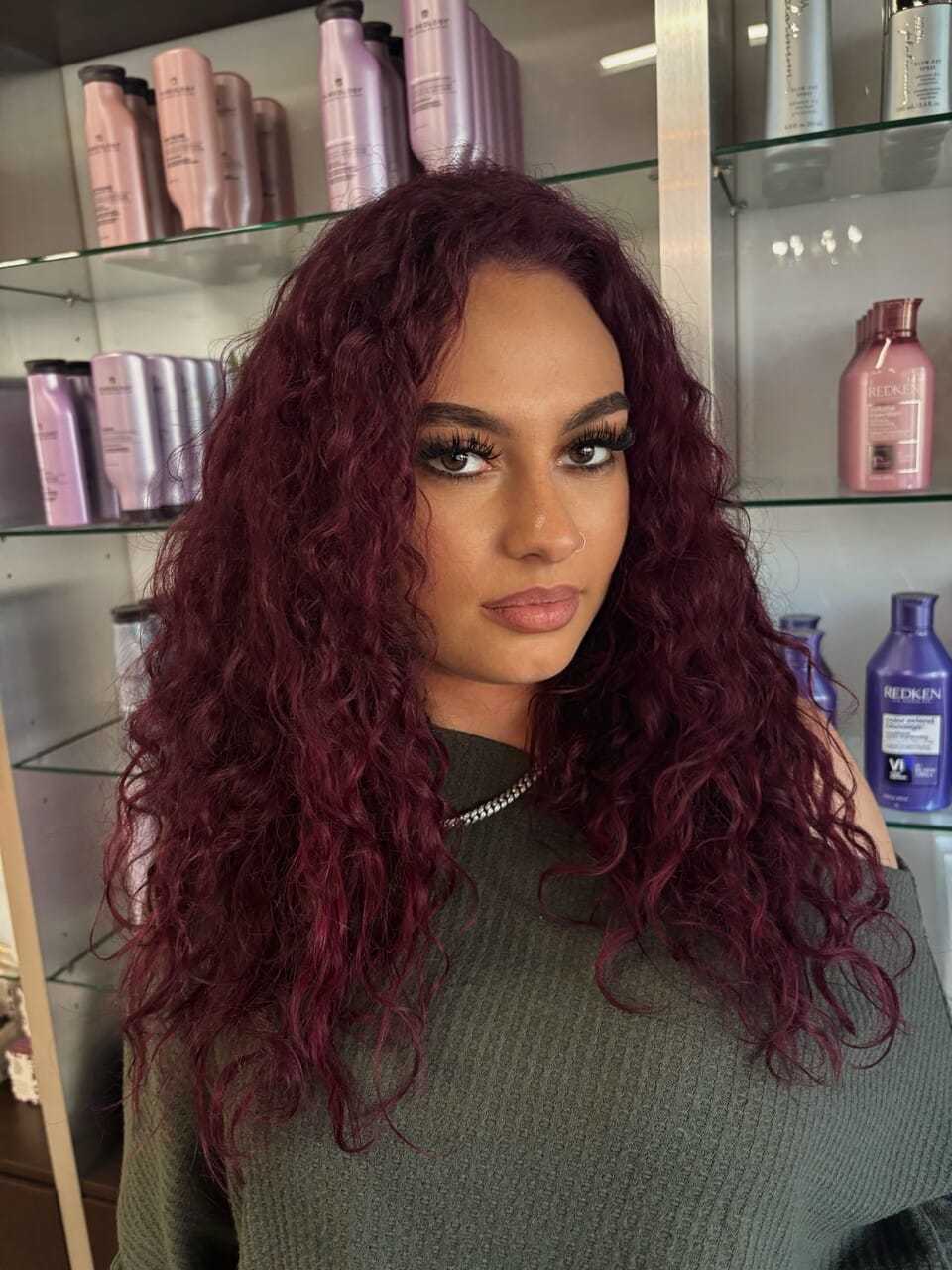 Woman with curly burgundy hair posing in front of salon products on shelves.